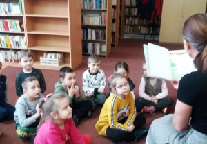 Dzieci siedzą na dywanie, pani bibliotekarka czyta im książkę.