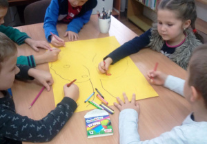 Dzieci siedzą przy stoliku, na którym leży arkusz żółtego papieru. Ich zadaniem jest narysowanie jabłoni.