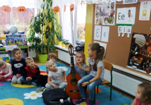 Dzieci siedzą w kręgu na dywanie, pod tablicą do zajęć na krzesełku siedzi Alicja, która prezentuje dzieciom swoją gitarę.