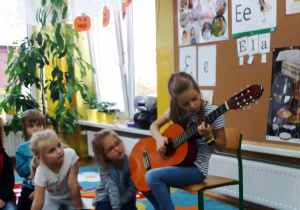 Grupka dzieci z uwagą przyglądają się grającej na gitarze dziewczynce. W tle tablica z ilustracjami, po lewej stronie duży stojący na podłodze kwiat.
