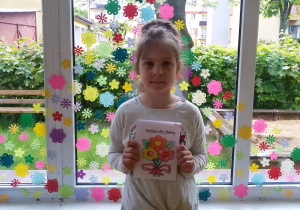 Widok na dziewczynkę stojącą na tle okna wyklejonego kolorowymi kwiatuszkami. Oliwia trzyma w rękach laurkę z napisem „Kwiaty dla Mamy. Pod napisem bukiet z trzech kwiatów, związanych czerwoną kokardą.