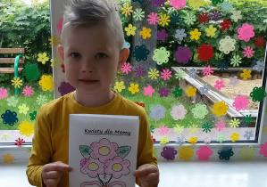 Widok na chłopca stojącego na tle ozdobionego kwiatkami okna. Filip trzyma w rękach laurkę z napisem „Kwiaty dla Mamy. Pod napisem bukiet z trzech różowych kwiatów, związanych czerwoną kokardą.