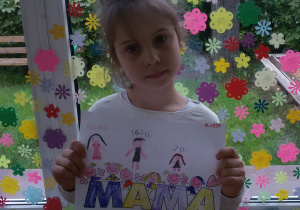 Widok na dziewczynkę stojącą na tle okna wyklejonego kolorowymi kwiatuszkami. Oliwia trzyma w rękach rysunek z napisem MAMA, litery M mają granatowy kolor, A żółty. Nad napisem znajdują się postacie dzieci nad nimi Oliwia narysowała mamę, tatę i siebie. Powyżej napisała: mama, tata, ja. W prawym górnym rogu napisała swoje imię.