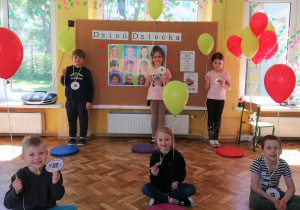 Sześcioro dzieci z balonikami. W tle tablica z napisem Dzień Dziecka i obrazkami przedstawiajacymi dzieci.