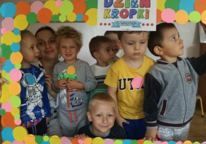 Dzieci wspólnie z nauczycielką pozują do zdjęcia stojąc za ramką ozdobioną różnokolorowymi kropkami.