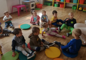 Grupa dzieci siedzi na kolorowych krążkach, przed nimi na krześle siedzi nauczycielka, która czyta im bajkę.