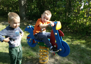 Widok na dwóch chłopców bawiących się na placu zabaw, jeden z nich siedzi na bujaku, a drugi stoi obok.