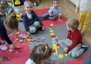 Dzieci siedzą na dywanie i prezentują ułożony z płaskich figur geometrycznych pociąg.