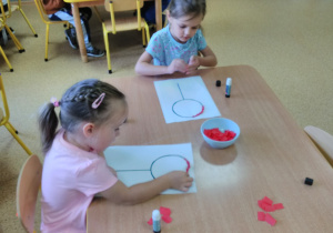 Widok na dwie dziewczynki siedzące przy stoliku. Na stoliku w miseczce są kawałki bibuły w kolorze czerwonym oraz kartki z narysowanym wzorem „lizak policjanta”, który składa się z koła i linii pionowej. Dzieci naklejają bibułki po śladzie wzoru.