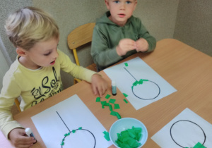 Dwóch chłopców siedzi przy stoliku, na którym w miseczce są kawałki bibuły w kolorze zielonym oraz kartki z narysowanym wzorem „lizak policjanta”, który składa się z koła i linii pionowej. Dzieci naklejają bibułki po śladzie wzoru.