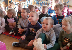 Widok na grupę roześmianych dzieci, które siedzą na dywanie ze swoimi misiami.