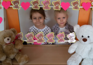 Widok na dwie dziewczynki, które stoją za ramką ozdobioną misiami i różowymi serduszkami.