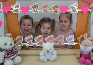 Widok na trzy dziewczynki, które pozują do zdjęcia stojąc za ramką ozdobioną misiami i różowymi serduszkami.