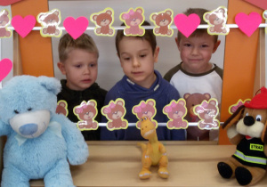 Widok na trzech chłopców, którzy stoją za ramką ozdobioną misiami oraz różowymi serduszkami i pozują do zdjęcia.