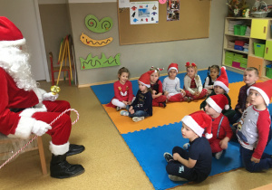 Dzieci siedzą na macie i słuchają z uwagą słów Św. Mikołaja, który siedzi przed nimi na krześle.
