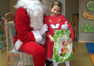 Widok na siedzącego na krześle Św. Mikołaja i dziewczynkę, która trzyma w rękach prezent.