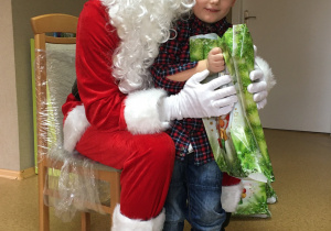 Widok na siedzącego na krześle Św. Mikołaja i stojącego przy nim chłopca, który trzyma w rękach prezent.