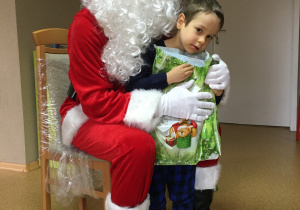 Widok na siedzącego na krześle Św. Mikołaja i stojącego przy nim chłopca, który trzyma w rękach prezent.