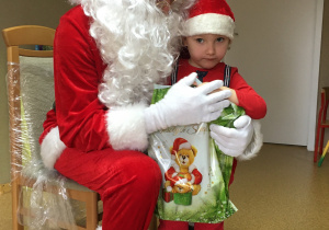 Widok na siedzącego na krześle Św. Mikołaja i stojące przy nim dziecko, które trzyma w rękach prezent.