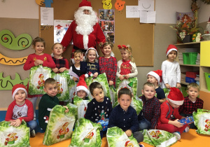 Zdjęcie grupowe. Grupa uśmiechniętych dzieci trzymających prezenty, za nimi stoi Św. Mikołaj. W tle widać tablicę korkową.