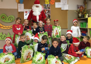 Zdjęcie grupowe. Grupa uśmiechniętych dzieci trzymających prezenty, za nimi stoi Św. Mikołaj. W tle widać tablicę korkową.