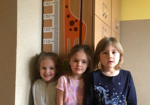 Trzy dziewczynki ustawiły się przy szafie, na której naklejona jest żyrafa z miarą w cm.