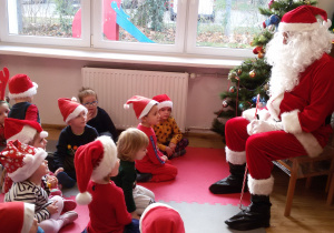 Widok na salę przedszkolną i dzieci siedzące na dywanie, przed nimi na krześle siedzi Święty Mikołaj. W tle okno, przez które widać ogród przedszkolny.