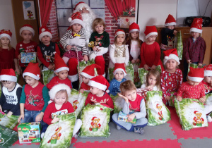 Dzieci w mikołajkowych czapeczkach z prezentami w rękach pozują do zdjęcia grupowego ze Świętym Mikołajem.