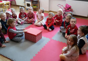 Dzieci siedzą w kręgu na dywanie, w środku siedzi chłopiec, który trzyma ręce na czerwonym pudle.