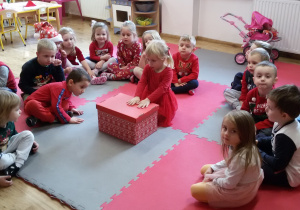 Dzieci siedzą w kręgu na dywanie, w środku siedzi dziewczynka, która trzyma dłonie na czerwonym pudle.