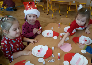 Widok na siedzące przy stoliku dzieci, które sklejają gotowe elementy tworząc głowę Świętego Mikołaja