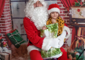 Dziecko siedzi na kolanach u Świętego Mikołaj. W tle świąteczna dekoracja.