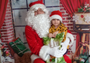 Dziecko siedzi na kolanach u Świętego Mikołaj. W tle świąteczna dekoracja.