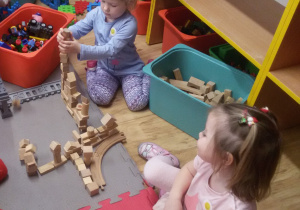 Widok na dwie dziewczynki, które wznoszą budowlę z drewnianych klocków.