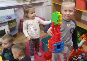 Widok na grupę bawiących się zabawkami dzieci. Jakub prezentuje wykonaną przez siebie budowlę z klocków.