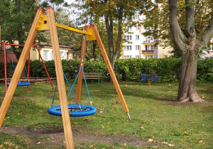 widok na ogródek przedszkolny, z przodu jest huśtawka, tak zwane bocianie gnazdo, dalej rośnie duże drzewo, z lewej strony widać fragment budynku przedszkola, huśtawkę oraz piaskownicę.